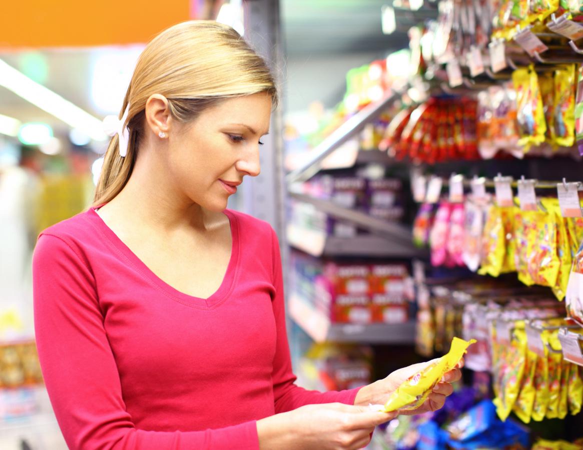 A woman wearing a pink shirt reads the ingredient label of a yellow package of candy in a grocery store.