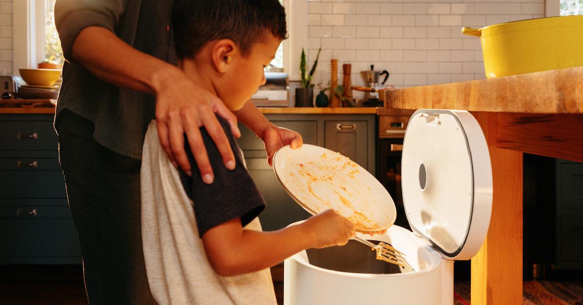 A parent helps a child scrape food scraps from a plate into a white Mill bin.