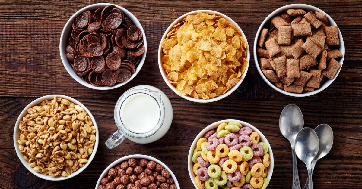 Six bowls of ultra-processed, sugary cereal on a table with a cup of milk and three spoons. 