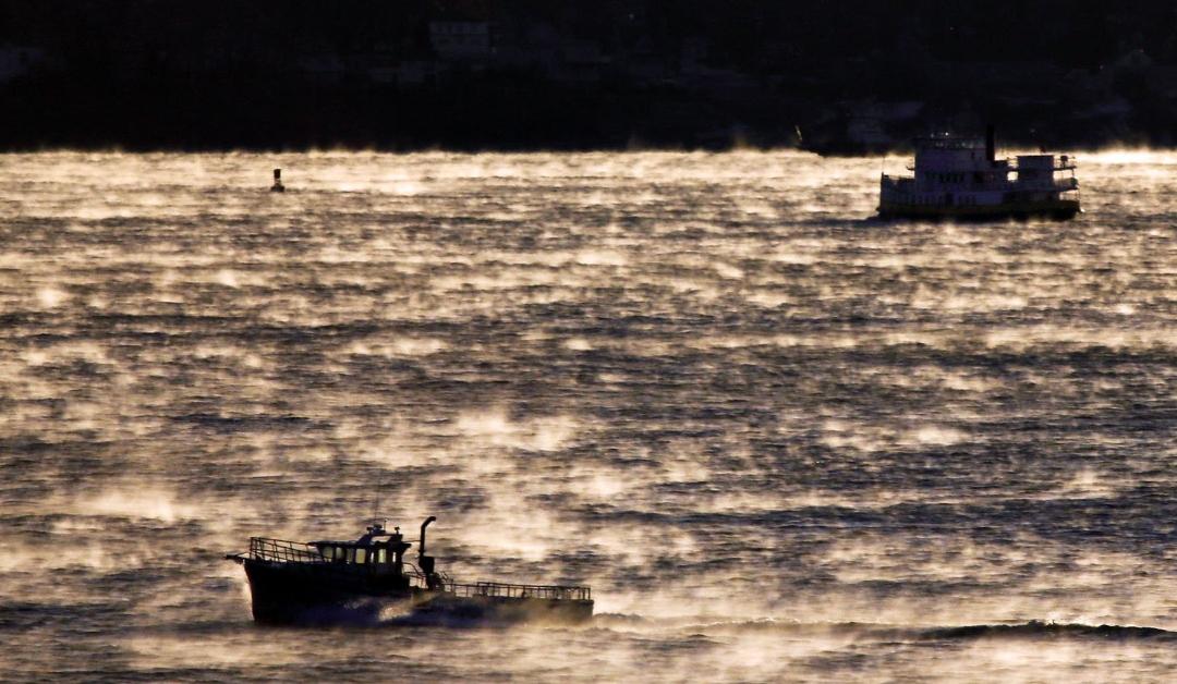 Two boats spotted on open, sunlit water through a thick haze of Arctic sea smoke