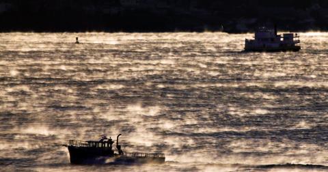 Two boats spotted on open, sunlit water through a thick haze of Arctic sea smoke