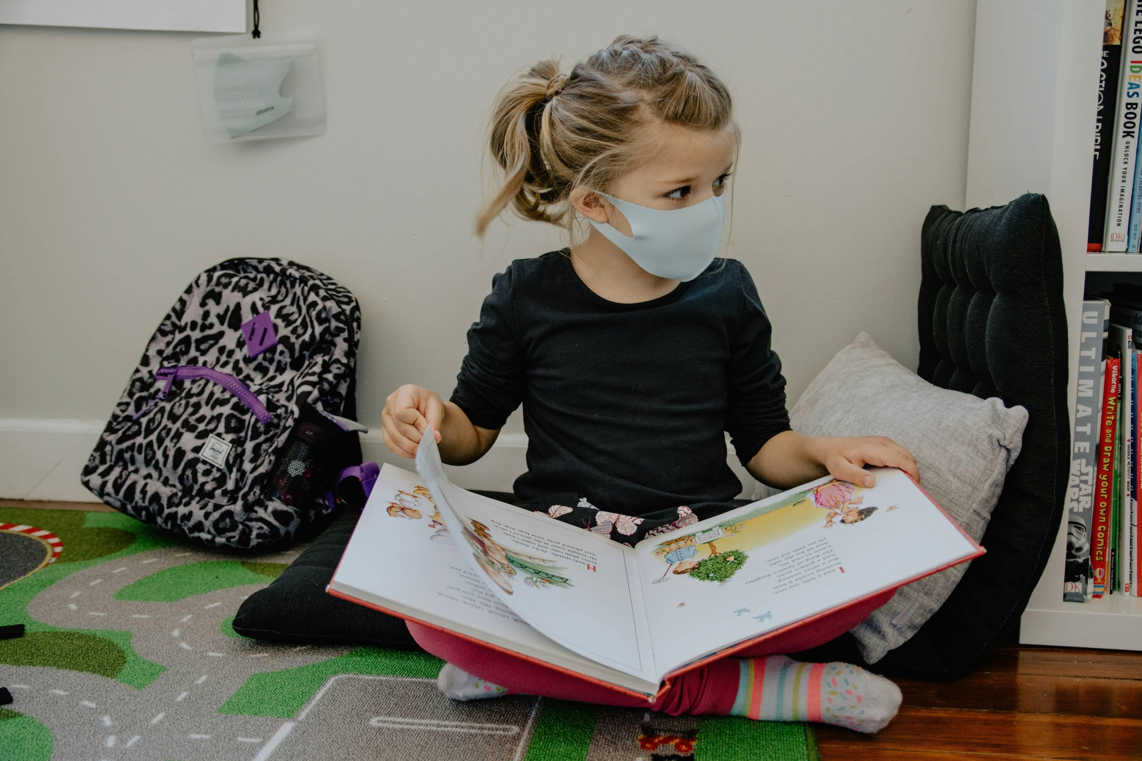 A young child sits on the floor in school while wearing a face mask and reading a book.