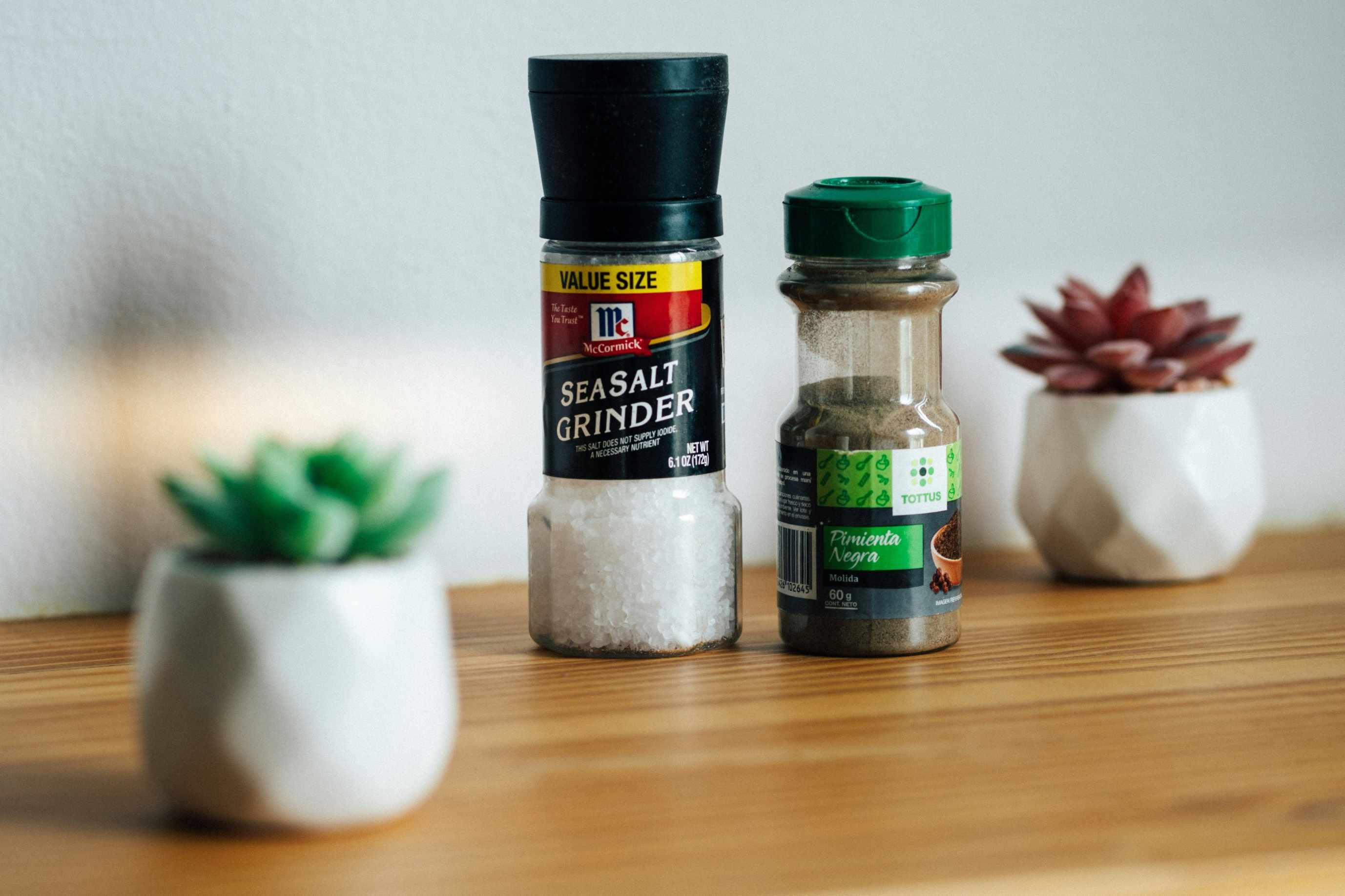 A picture of a sea salt grinder next to a black pepper container and two succulents.