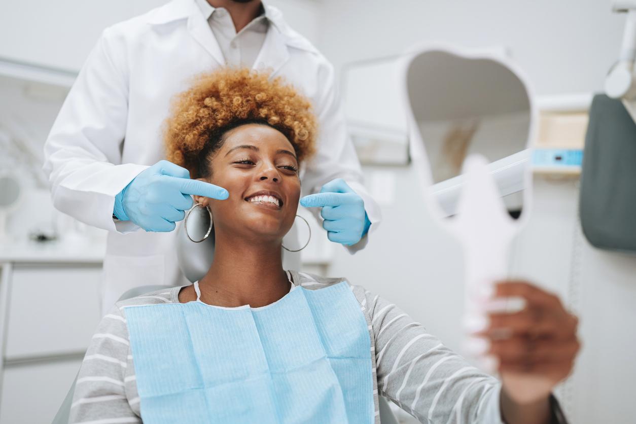 A smiling woman with a blue bib lays in a dentist chair while the dentist touches the sides of her mouth with gloved hands.