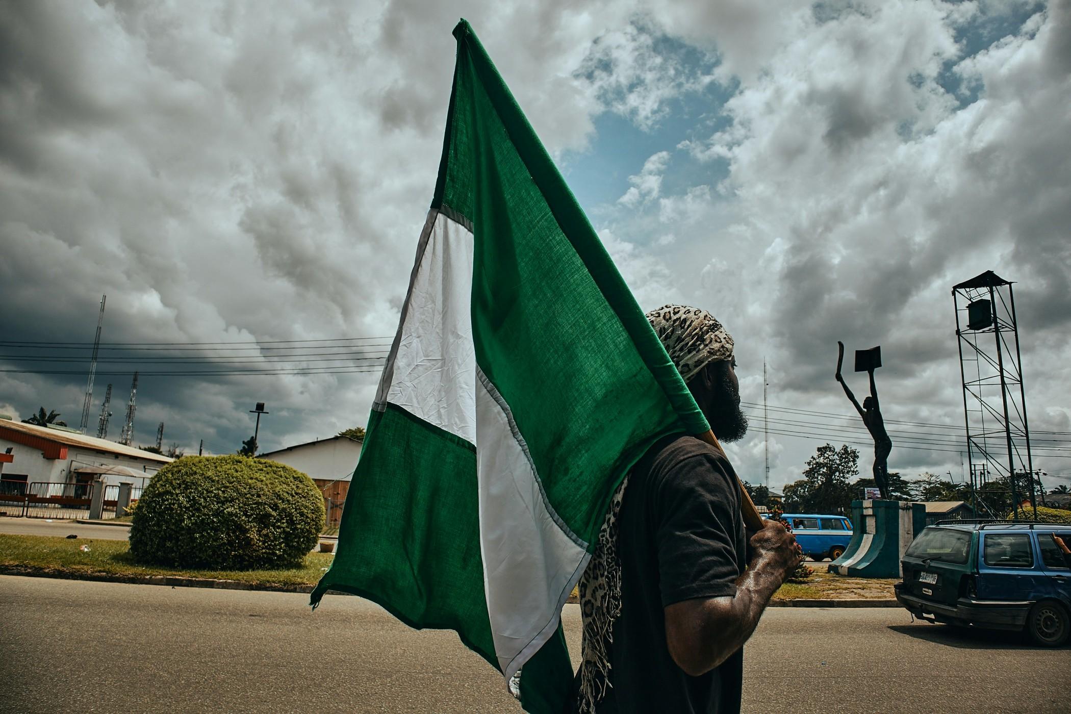 A Nigerian is pictured from the side while standing in the street holding the flag of Nigeria behind him.