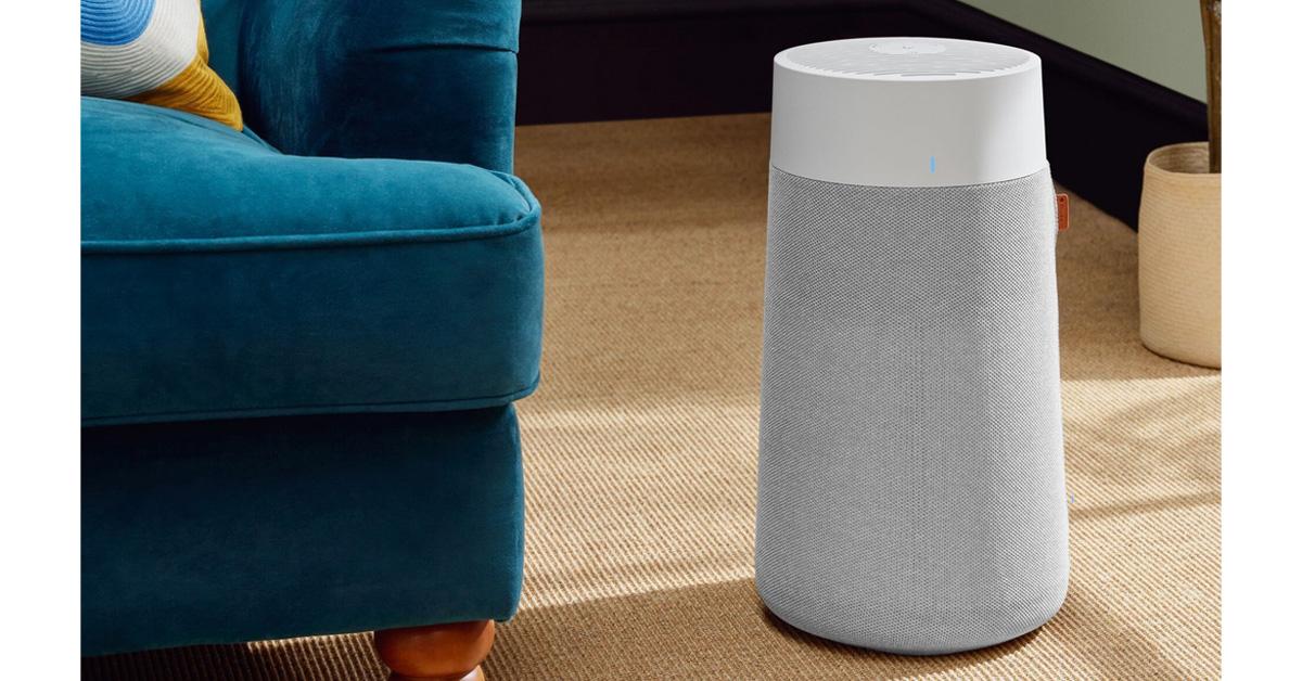 an air purifier on the floor of a room with a rug, chair, and plant