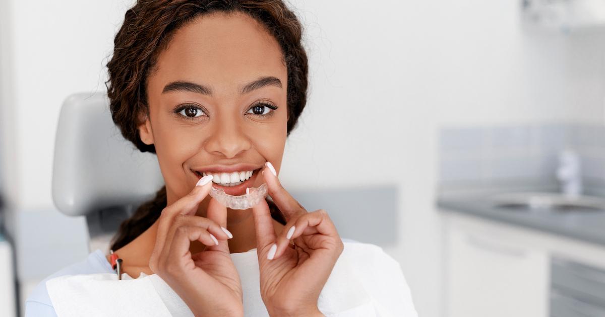 Smiling black girl holding invisible teeth aligner in dentist chair