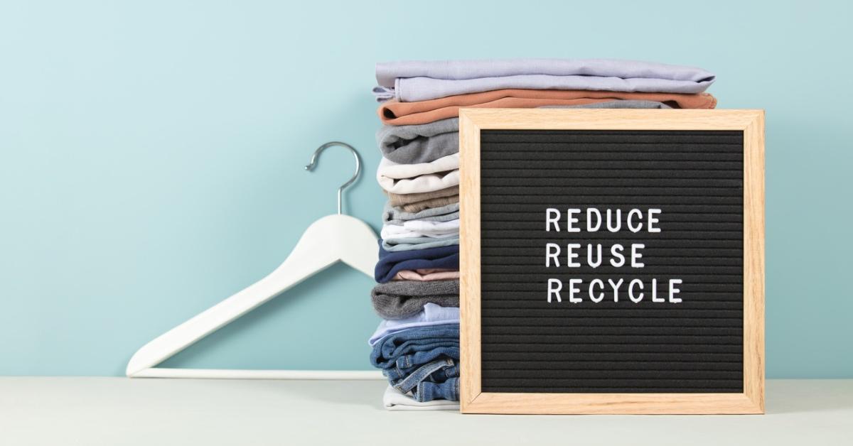 Folded pieces of clothing against a blue background with a board that says Reduce, Reuse, Recycle.