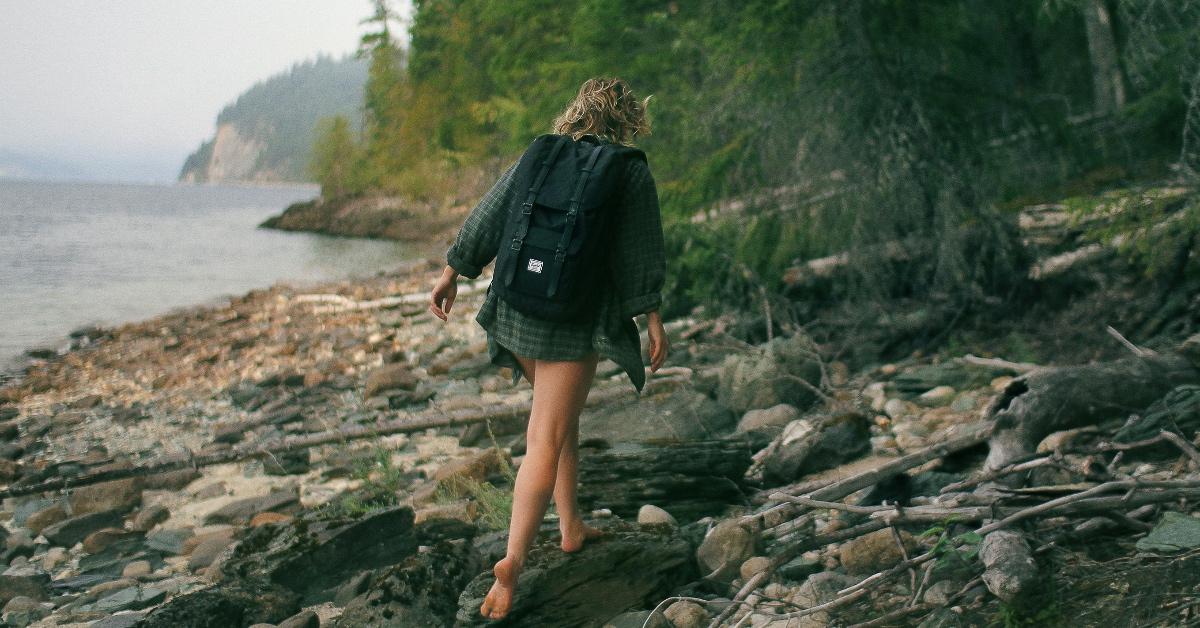 A woman with a backpack walking barefoot across a rocky coast. 