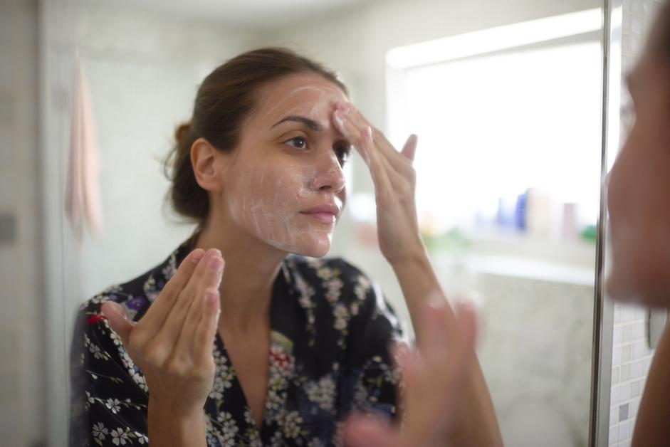 A woman looks in the mirror while applying face cleanser to her face. 