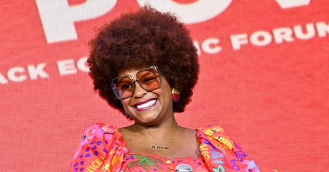 Tabitha Brown smiles in front of a red background, wearing sunglasses and a colorful top.
