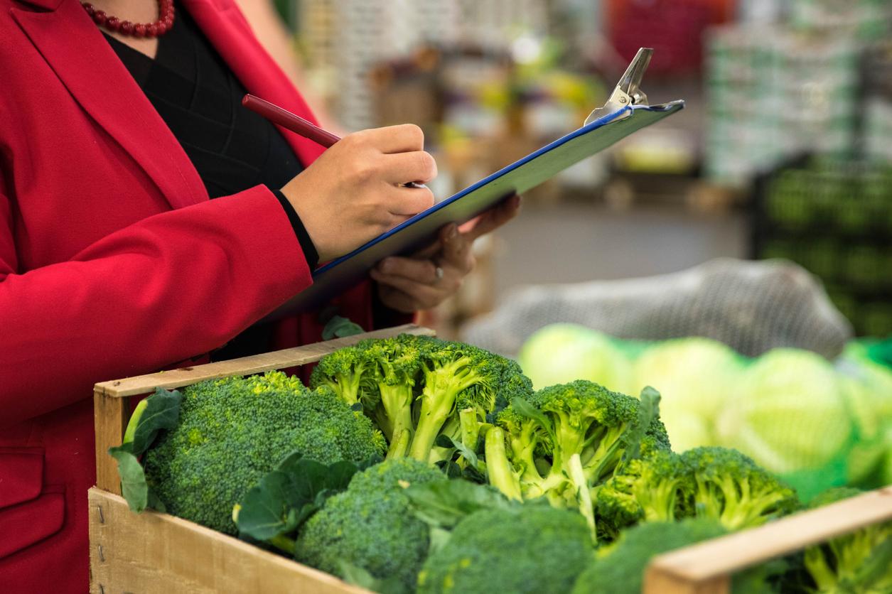 An employee in a red suit writes on a clipboard after inspecting broccoli for freshness.