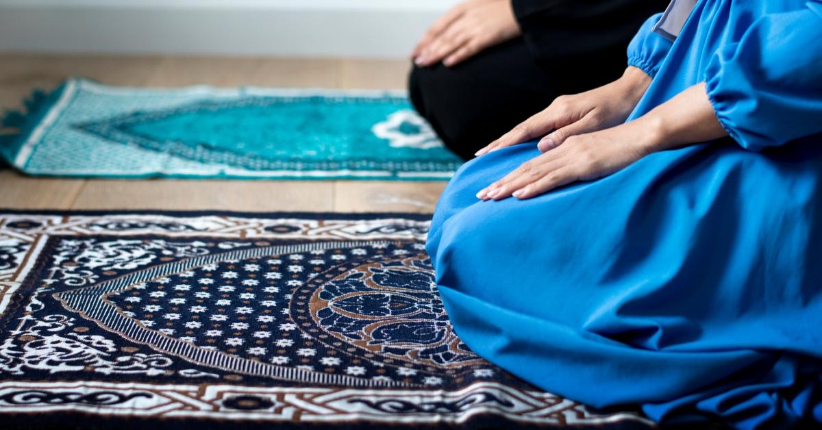 Two Muslims wearing black and blue clothing kneel on prayer rugs with their hands on their legs. 