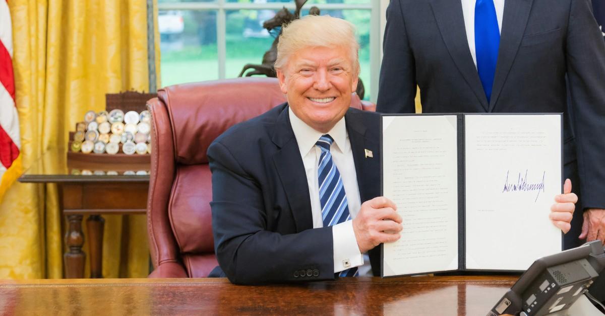 Donald Trump smiles while holding up a folder with his signature on it