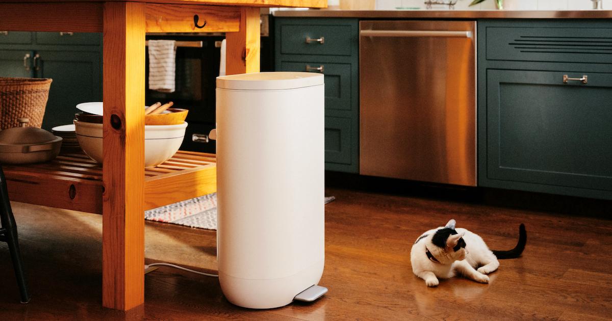 A white Mill compost bin sits on the floor of a kitchen, next to a kitchen island, and a cat rests on the floor.