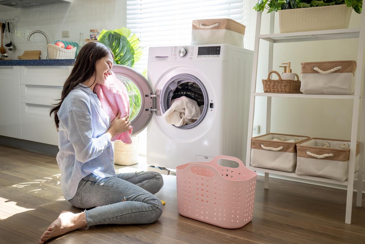 A smiling woman sits by an open dryer with her eyes closed while smelling a freshly laundered pink blanket in her laundry room.