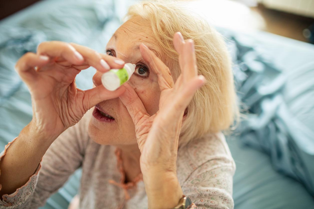 An older adult woman in bed holds her left eye open with one hand while squeezing eye drops into her eyes with another hand.