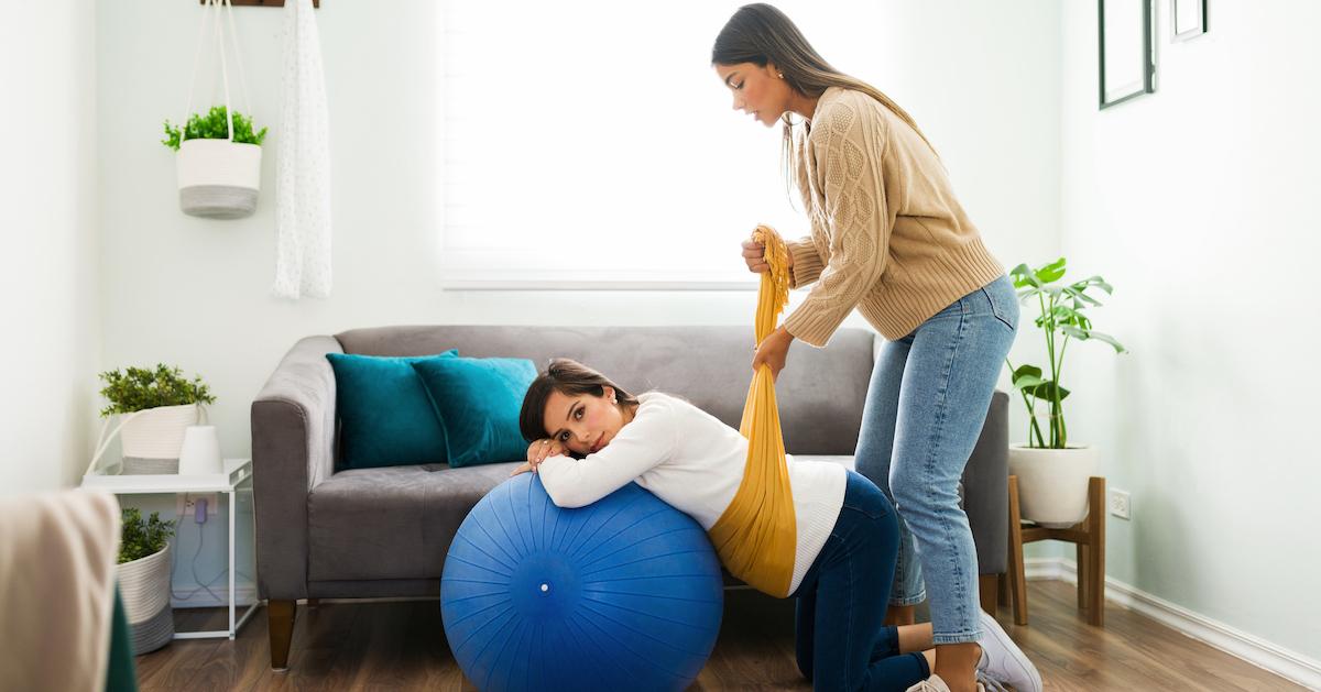 A pregnant woman lays on an exercise ball, and a doula gently lifts her belly with a scarf.