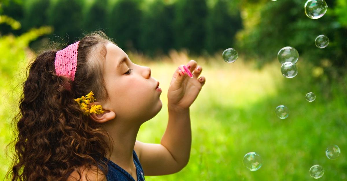 Little girl with brown hair blows bubbles outside in a grassy lawn.