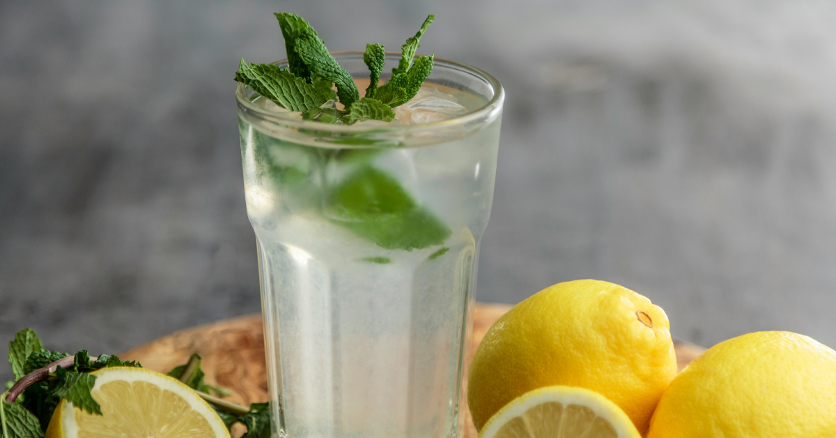 A glass of cold water that has been infused with lemon and mint, surrounded by cut and uncut lemons