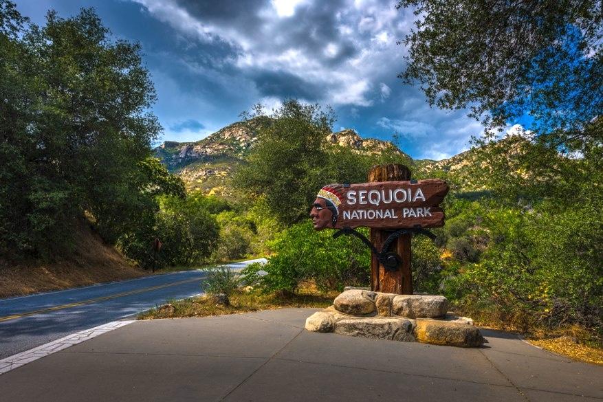 The entrance to Sequoia National Park is depicted against a backdrop of trees and a mountains and a cloudy sky.