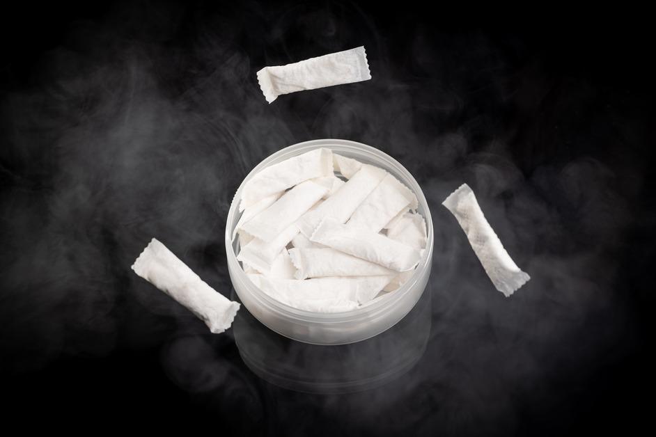 A container of nicotine pouches with smoke around it sits on a black background. 