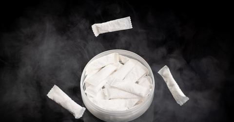 A container of nicotine pouches with smoke around it sits on a black background.