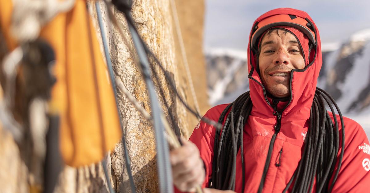 Alex Honnold has a bloody nose, and wears an orange helmet and red jacket, holding ropes on mountain, smiling at the camera.