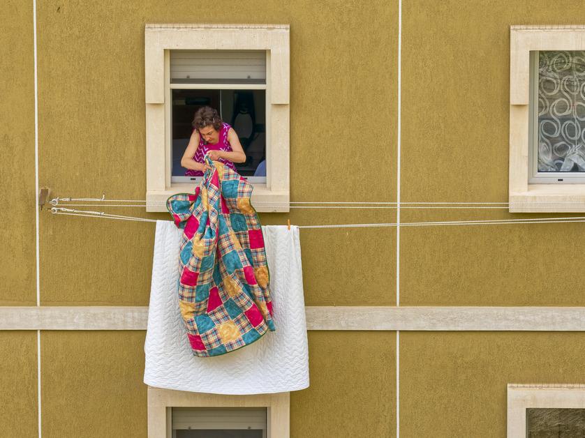 A woman wearing a pink dress holds out a quilt from a yellow building. 