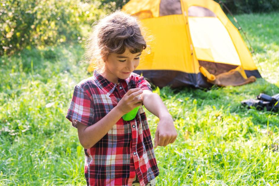 A young girl in a plaid shirt stands outside with a yellow tent in the background and puts on bug spray.