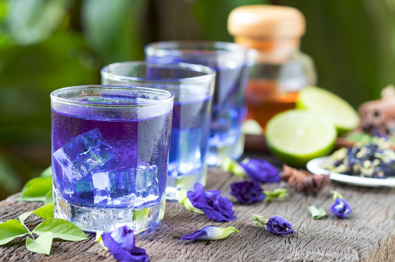 A line of glass of iced butterfly pea flower tea are pictured atop a table with flowers and lime wedges beside them.