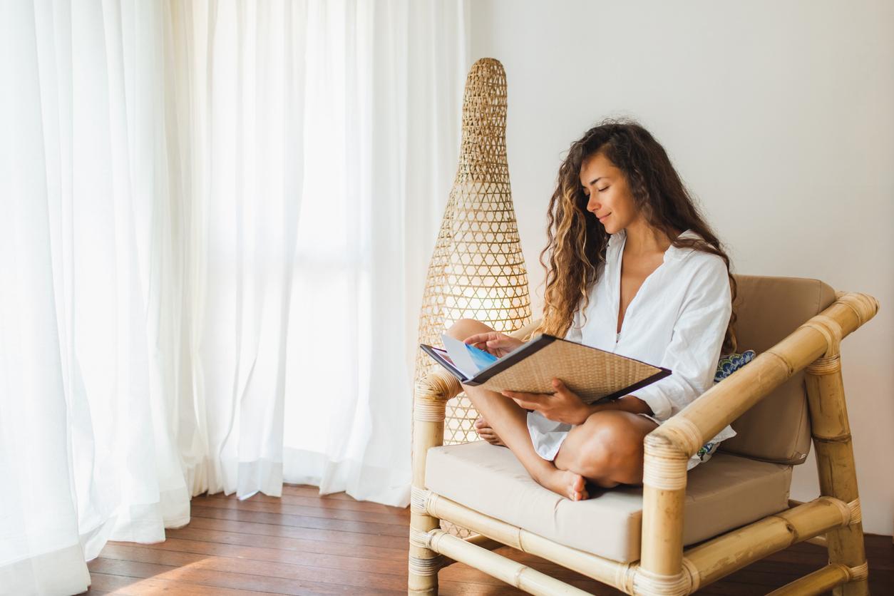 A smiling woman reads a book while sitting in a bamboo chair in her living room.