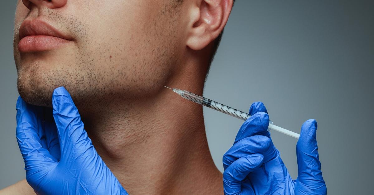 A pair of hands wearing blue gloves holds an injection syringe to a man's jawline. 