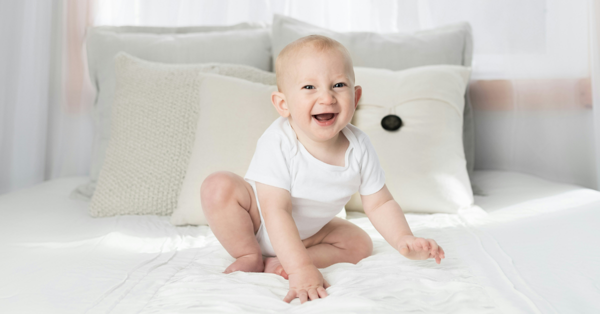 An infant sits on a bed smiling