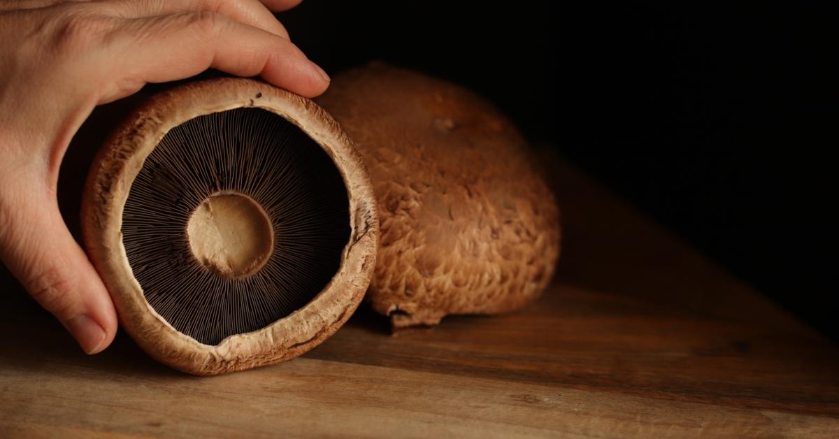 A person's hand holds up the inside of a portobello mushroom on a wooden surface with other mushrooms in the background. 