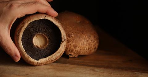 A person's hand holds up the inside of a portobello mushroom on a wooden surface with other mushrooms in the background.
