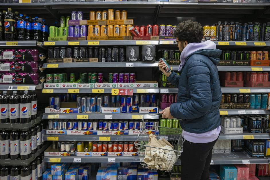 A man wearing a blue puffer coat stands in front of the energy drink isle at the store while holding up a drink and looking at the label. 