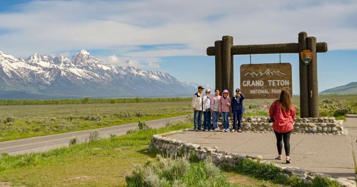 Group of tourists posing in front of a sign at the entrance to the Grand Teton National Park (Representative Cover Image Source: Getty Images | Ceri Breeze)