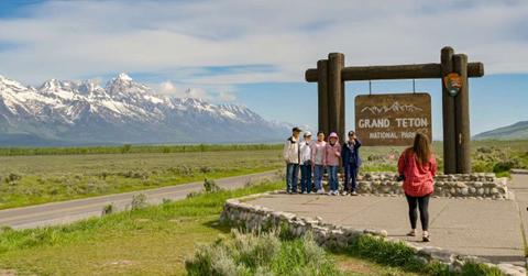 Group of tourists posing in front of a sign at the entrance to the Grand Teton National Park (Representative Cover Image Source: Getty Images | Ceri Breeze)