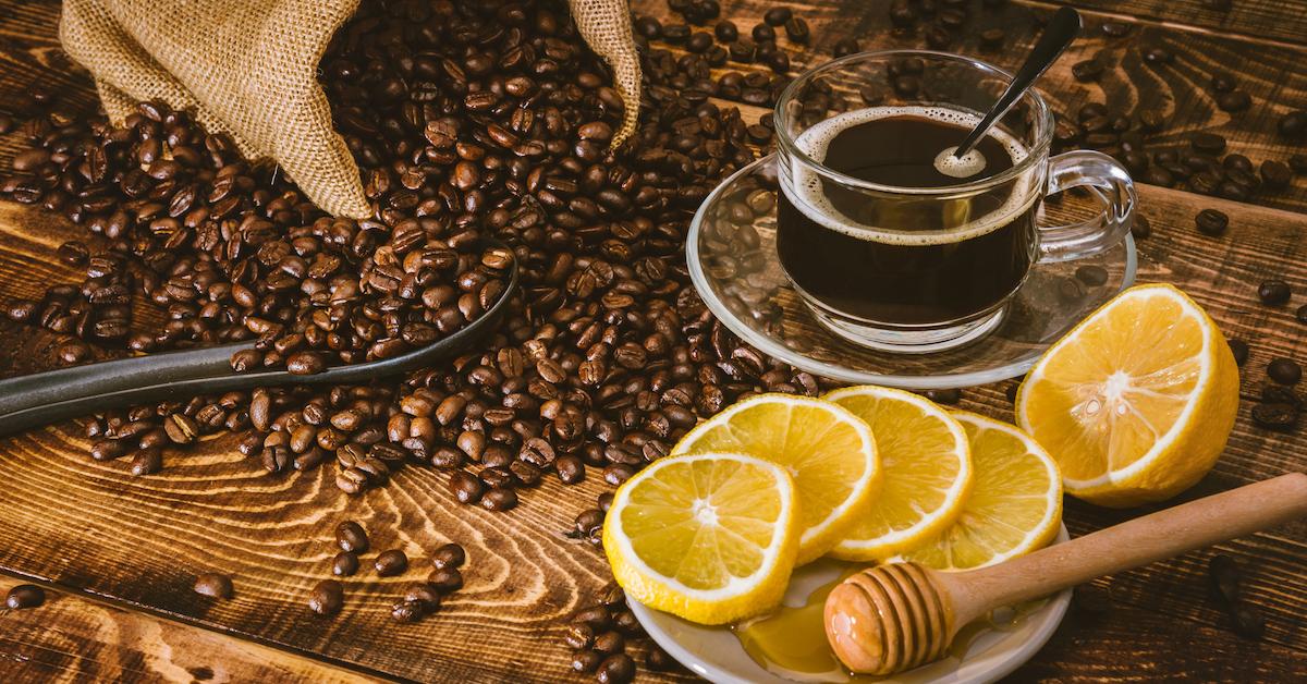 Coffee beans, a cup of coffee, a plate of lemon slices, and a honey dipper on a wooden table.