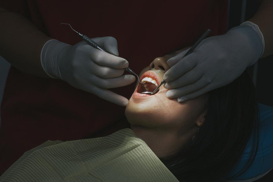 A woman sits in a chair at the dentist with a light shining on her mouth while the dentist uses tools on her teeth. 