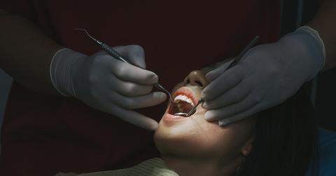 A woman sits in a chair at the dentist with a light shining on her mouth while the dentist uses tools on her teeth.