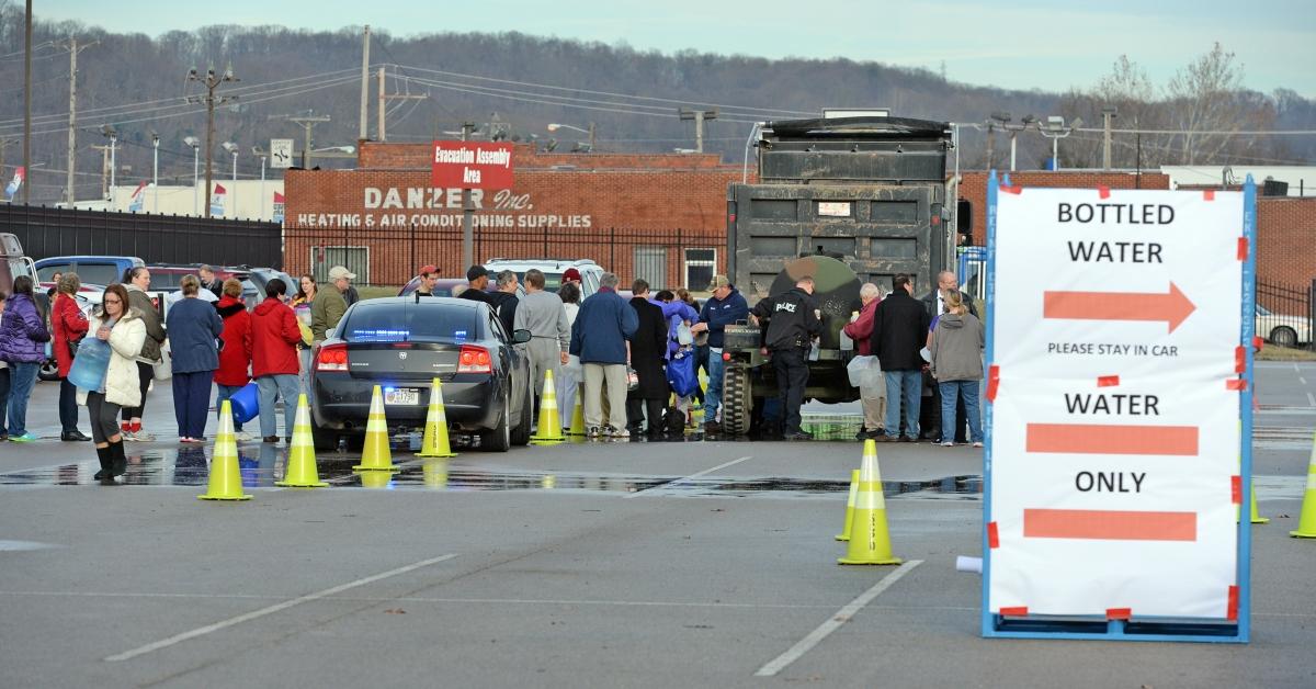 In a parking lot, people line up to fill water jugs.