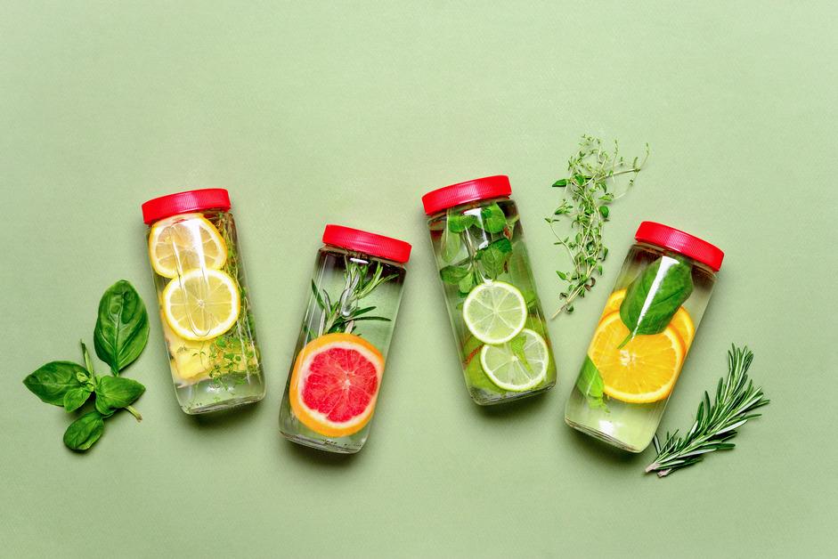 Four jars of water laying on a green background filled with lemon, grapefruit, orange, lime slices, and herbs. 