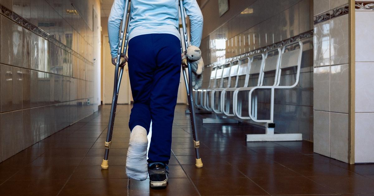 Girl walks down hallway on crutches with broken foot holding teddy bear.