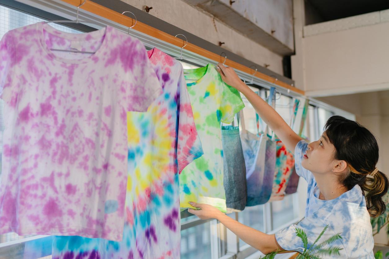 Person touching and looking one tie-dye shirt that's hanging up among other tie-dye shirts and tote bags.