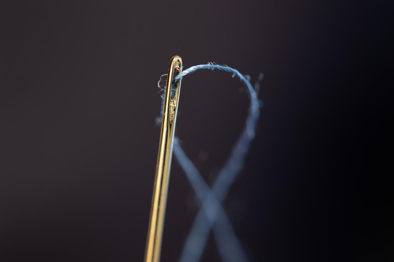 Very close up view of blue thread running through the eye of a sewing needle.