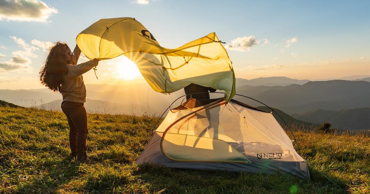 Woman fans a tarp next to a tent, in front of a sunset on a mountain