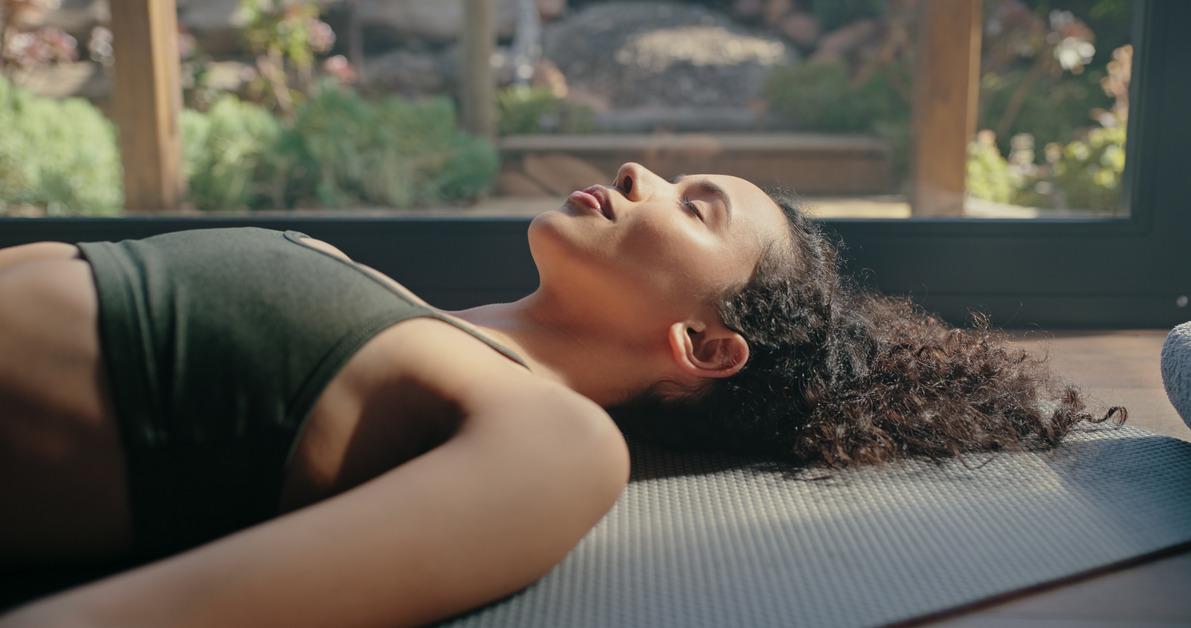 Woman with curly hair and a green sports bra lies down on a yoga mat in front of a window with her eyes closed. 