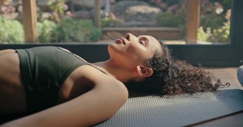 Woman with curly hair and a green sports bra lies down on a yoga mat in front of a window with her eyes closed.